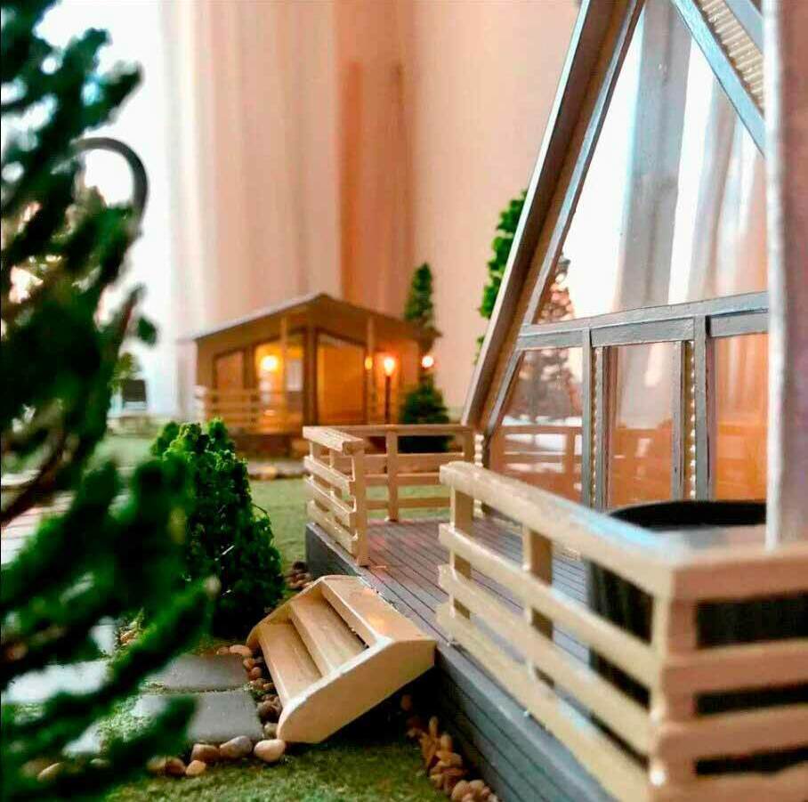 Миниатюрная модель загородного дома из интерьерного конструктора румбокса. На переднем плане — деревянная терраса с перилами, ведущая к входу в дом. Рядом с террасой расположены декоративные камни и миниатюрные кустарники, создающие ощущение ухоженного сада. Дом выполнен в современном стиле с большими панорамными окнами, пропускающими много света. На заднем плане видна ещё одна постройка, напоминающая беседку или хозяйственное помещение, с тёплым освещением внутри. Вокруг моделей — имитация зелёных насаждений, подчёркивающая уют и гармонию композиции. Модель идеально подходит для творчества, коллекционирования и украшения интерьера, позволяет воссоздать кукольный мир в масштабе 1:43.