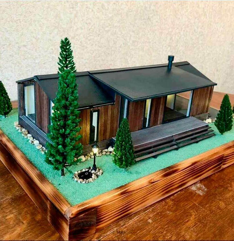 Макет современного загородного дома с ландшафтным дизайном: детализированная модель коттеджа с террасой, хвойными деревьями и декоративной подсветкой для архитектурных презентаций Масштабная модель загородного дома в современном стиле: одноэтажное здание с большими панорамными окнами, тёмной крышей и деревянной отделкой фасада. Перед домом — просторная терраса с лестницей, ведущая к ухоженному участку. Ландшафтный дизайн включает миниатюрные хвойные деревья, аккуратно выложенные камни вдоль фундамента, зелёное покрытие, имитирующее газон. Особое внимание уделено элементам освещения — установлен декоративный фонарь. Модель демонстрирует гармоничное сочетание архитектуры и озеленения, идеально подходит для визуализации проектов частных домов и планирования придомовой территории.