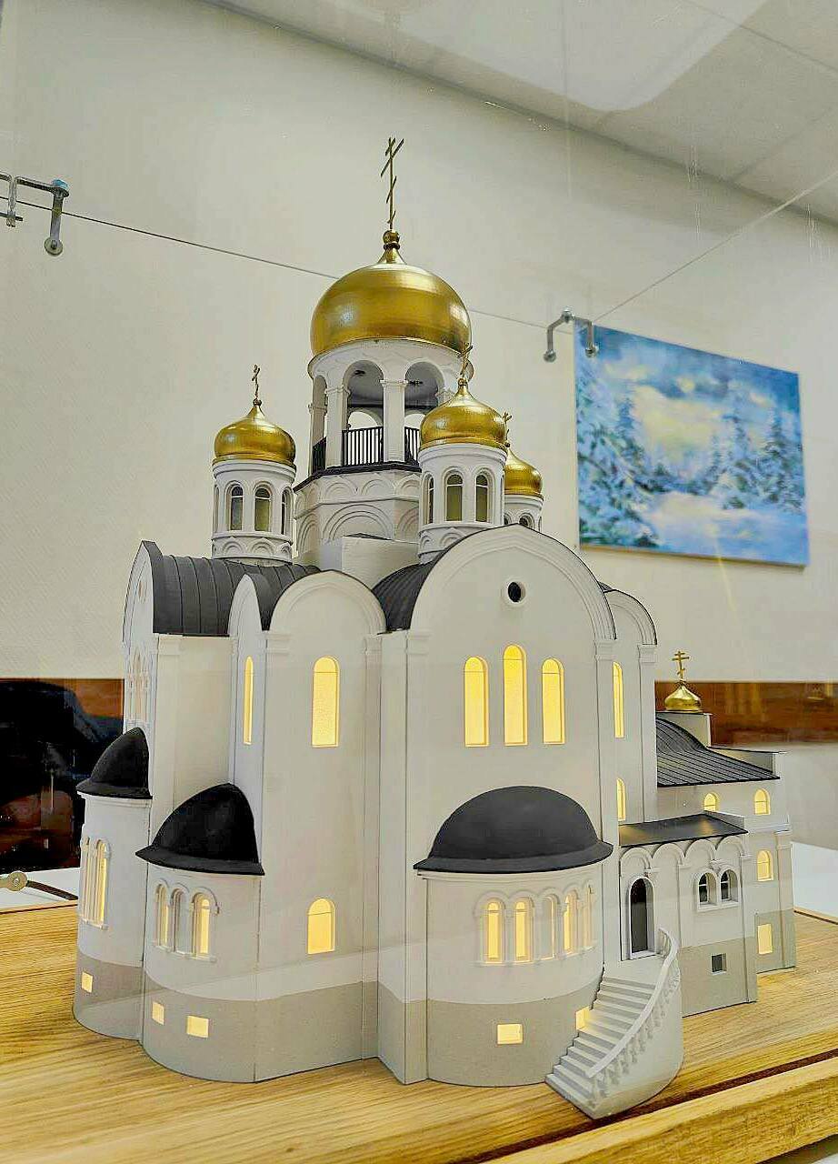 Макет православного храма с золотыми куполами: детализированная архитектурная модель для выставок и презентаций Детализированный масштабный макет православного храма: белокаменные стены, арочные окна и порталы, чёрные кровли, золотые купола с крестами; в проёмах — тёплая подсветка, у входа — тёмная площадка с крапчатой фактурой.