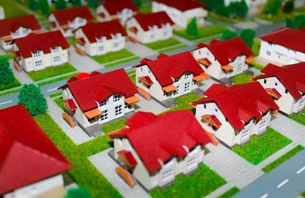  Детальный макет коттеджного посёлка в масштабе: аккуратно выстроенные ряды двухэтажных домов с выразительными красными крышами и светлыми фасадами. Каждый дом окружён ухоженным зелёным газоном, рядом расположены миниатюрные деревья и кустарники. Видны асфальтированные дороги и тротуары, чётко выделенные участки и подъездные пути. Модель демонстрирует сбалансированную планировку территории — гармоничное сочетание жилой застройки и зелёных зон. Отлично подходит для визуализации проектов загородной недвижимости, планирования коттеджной застройки, архитектурных презентаций перед инвесторами и потенциальными покупателями.