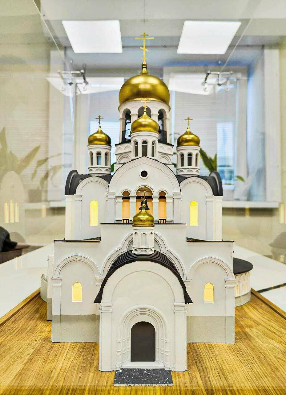 Макет православного храма с золотыми куполами: детализированная архитектурная модель для выставок и презентаций Детализированный масштабный макет православного храма: белокаменные стены, арочные окна и порталы, чёрные кровли, золотые купола с крестами; в проёмах — тёплая подсветка, у входа — тёмная площадка с крапчатой фактурой.