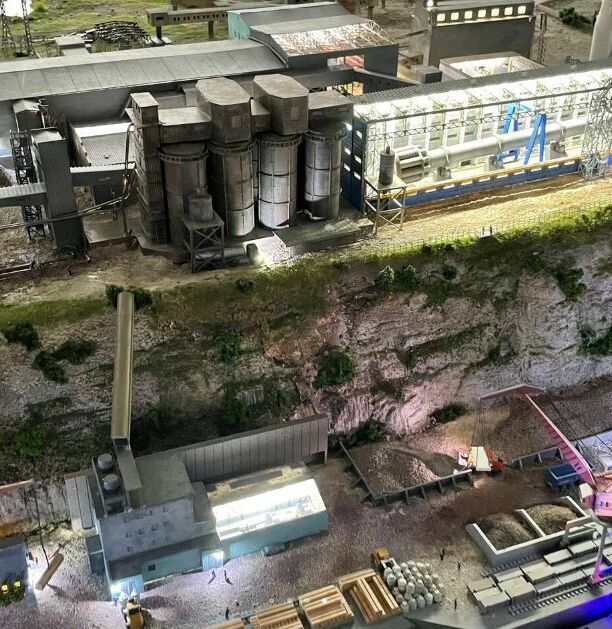 Детализированный 3D-масштабный макет промышленного предприятия / завода, расположенного на берегу водоёма. В композиции — крупный производственный корпус с конвейерными линиями и технологическим оборудованием, несколько вспомогательных цехов и складских помещений. Выделяются две высокие дымовые трубы, одна из которых окрашена в красно-белые полосы. Слева размещён грузовой порт с причалом, кранами для погрузки/разгрузки и пришвартованным судном. Видны транспортировочные пути, подъездные дороги, элементы ландшафтного дизайна (склоны, растительность). Макет демонстрирует полный цикл работы промышленного комплекса — от приёма сырья через производственные процессы до отгрузки готовой продукции. Реалистичная имитация инфраструктуры с высокой степенью детализации для использования в презентациях, планировании и визуализации проектов.