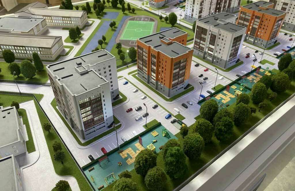 Детальный макет жилого комплекса, демонстрирующий планировку квартальной застройки: многоэтажные жилые дома с фасадами в современном стиле, благоустроенные дворы с детскими игровыми площадками, спортивная зона с футбольным полем, здания школы, парковочные места, зелёные насаждения и пешеходные дорожки. Визуализация комфортной городской среды с учётом всех элементов социальной инфраструктуры — образец комплексного подхода к проектированию жилых районов.