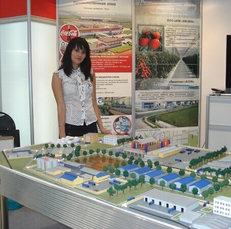 Масштабная модель агропромышленного комплекса, демонстрирующая планировку тепличного хозяйства с прилегающей инфраструктурой. В центре композиции — группа промышленных теплиц с прозрачными покрытиями, соединённых крытыми переходами. Рядом расположены складские помещения для хранения урожая, административный корпус с парковкой, зона погрузки/разгрузки с миниатюрными грузовиками. На модели видны элементы ландшафтного дизайна: аллеи, газоны, декоративные кустарники. Проработаны детали дорожной сети — основные и вспомогательные проезды, пешеходные дорожки. Также изображены инженерные коммуникации (миниатюрные линии электропередач, трубопроводы). Модель выполнена с высокой детализацией, позволяет наглядно оценить планировку комплекса, распределение функциональных зон и логистику перемещения продукции. Подходит для презентаций перед инвесторами, участия в отраслевых выставках, планирования застройки сельскохозяйственного предприятия.