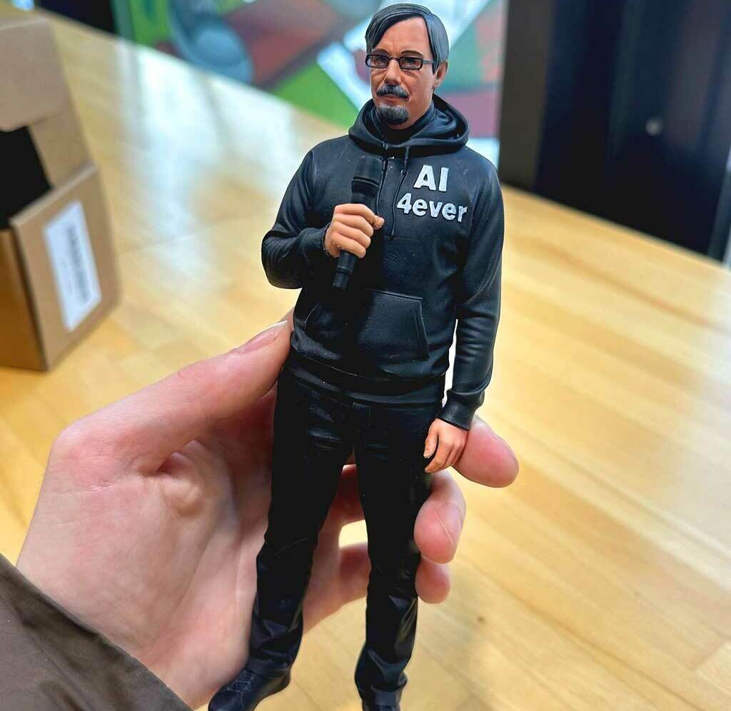 Реалистичная 3D-фигурка человека на заказ по фото: детализированная статуэтка с микрофоном, напечатанная на 3D-принтере. Кастомная модель в чёрной худи с надписью — идеальный сувенир для коллекционеров и поклонников публичных выступлений! 3D-фигурка мужчины, изготовленная по фотографии на 3D-принтере. Статуэтка изображает человека в расслабленной позе с микрофоном в руке. Одет в чёрную худи с надписью «AI ever», чёрные брюки, носит очки и аккуратную бороду. Высокая детализация: проработаны черты лица, текстура одежды, поза передаёт атмосферу публичного выступления. Фигурка отлично подойдёт для украшения офиса, в качестве оригинального подарка или элемента коллекции 3D-моделей. Пример персонализированной печати с сохранением индивидуальных особенностей.