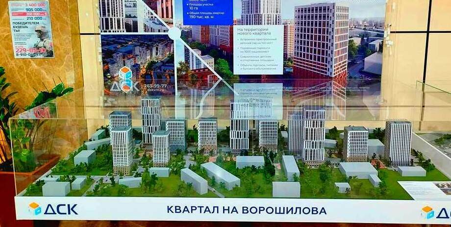 Макет жилого квартала с несколькими высотными зданиями, окруженными зелеными насаждениями. На заднем плане плакат с информацией о проекте: "Жилой квартал на Ворошилова", указаны параметры проекта, такие как население 60,000 человек, площадь участка 10 га, общая площадь квартир 190 тыс. кв. м.  На столе под прозрачным кожухом размещён масштабный макет застройки: группа многоквартирных жилых домов башенного и среднеэтажного типа, ориентированных кварталами. • Прорисованы внутриквартальные улицы/проезды, дворовые пространства, озеленение (деревья, аллеи), малые формы и площадки. • Здания разной высоты образуют композицию из нескольких доминант (высотных секций) и более низких корпусов по периметру. • Вокруг макета — информационный стенд с визуализациями и параметрами проекта, брендирован под «ДСК», заголовок «Жилой квартал на Ворошилова».