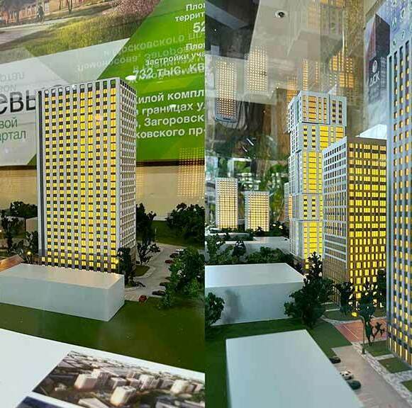 Архитектурный макет жилого комплекса с несколькими высотными зданиями. Здания имеют множество окон, подсвеченных изнутри, создавая эффект освещения. Вокруг зданий расположены небольшие деревья и кустарники, а также видны дороги и парковочные места. На заднем плане виден баннер с надписью "Яблоневый сад" и другой текст, связанный с проектом. 