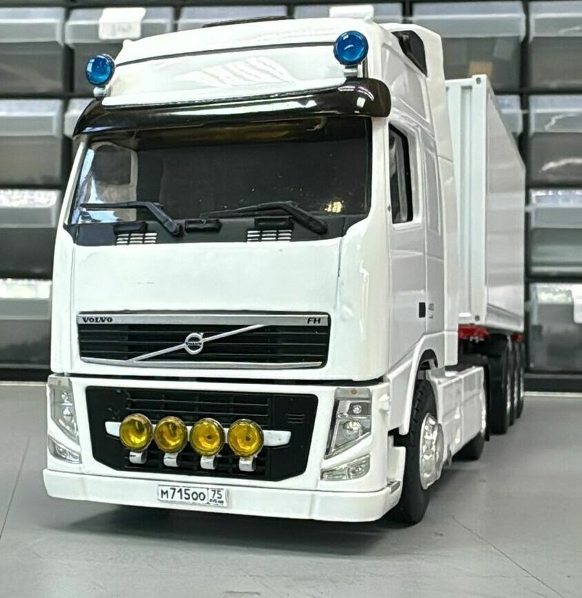 На фото показан стендовый макет магистрального автопоезда.  Техническое описание: • Тип ТС: седельный автопоезд европейской компоновки (кабover).   • Головное ТС: масштабная модель магистрального седельного тягача Volvo FH, колёсная формула ориентировочно 4×2.   • Надстройка: полуприцеп-фургон прямоугольного сечения (вероятно изотермический/контейнерного типа).   • Оснащение кабины: дополнительная блок-фара с четырьмя противотуманными/дальними фарами на переднем бампере, два синих проблесковых маяка на крыше, солнцезащитный козырёк над лобовым стеклом.  