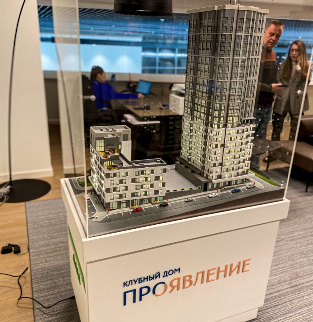 Макет здания, размещенная в прозрачной витрине. Модель представляет собой комплекс из двух зданий: одно высокое, многоэтажное, а другое более низкое. На подставке витрины написано "КЛУБНЫЙ ДОМ ПРОЯВЛЕНИЕ". 