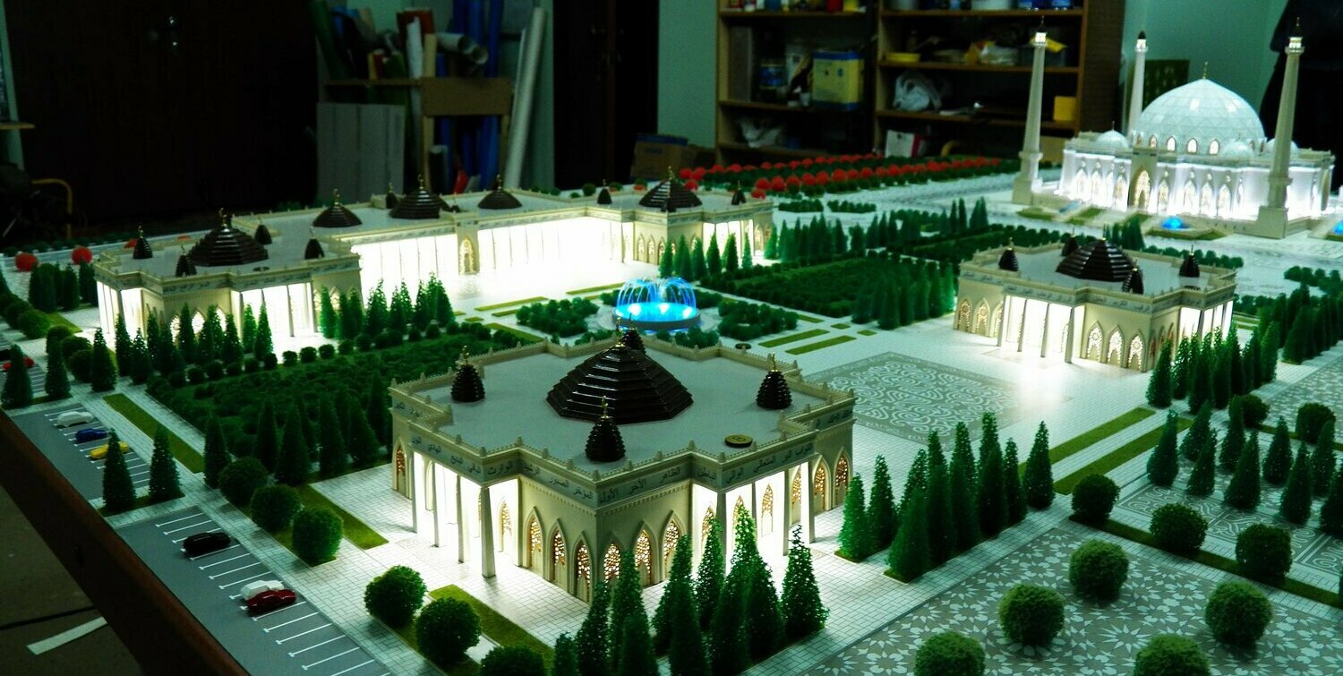 Детализированный макет мечети и окружающих павильонов: центральный купол, четыре высоких минарета, подсвеченные арочные входы, террасированные площади с орнаментальной плиткой, синие декоративные фонтаны, симметричные аллеи, миниатюрные деревья и парковки