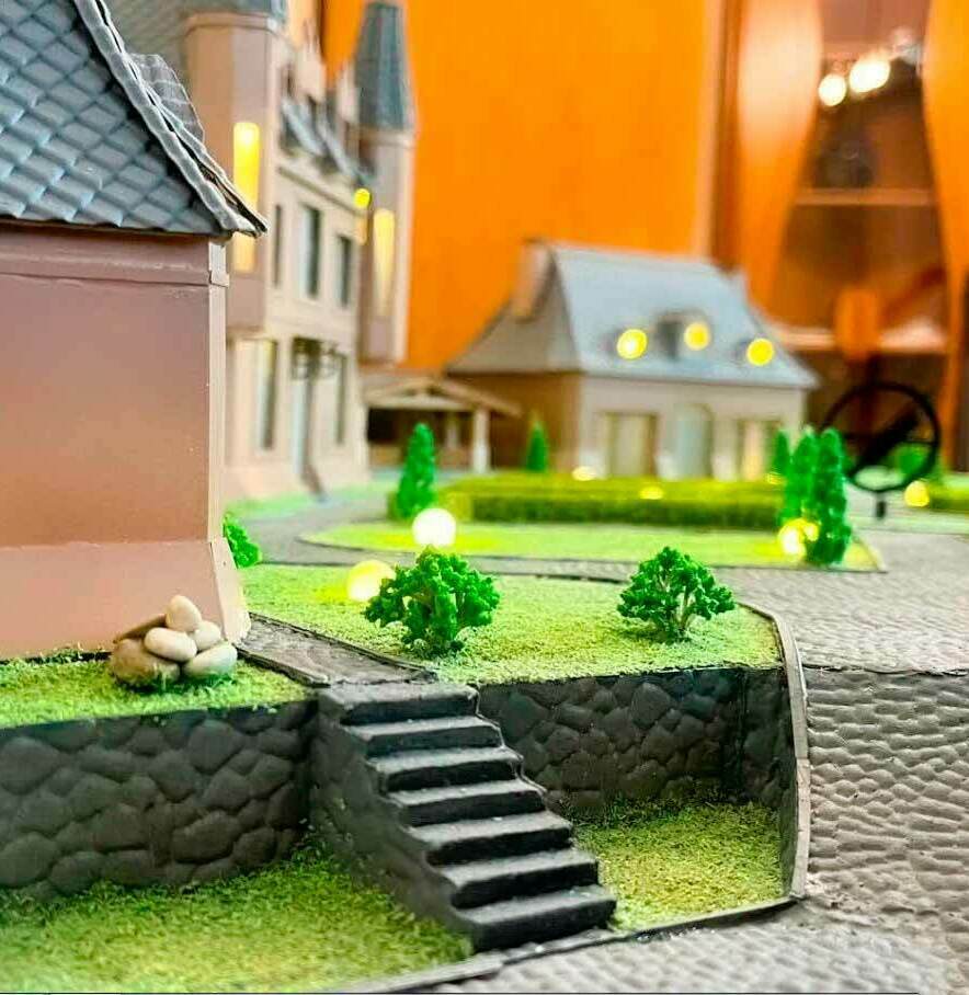 Детальный ландшафтный макет с архитектурными постройками: модель усадебной территории с подсветкой, лестницей, зелёными насаждениями и имитацией каменного ограждения — инструмент для презентации проектов загородной застройки Масштабная модель усадебной территории: на переднем плане — лестница, ведущая к участку с миниатюрными зелёными кустарниками и декоративными камнями. Видна имитация каменного ограждения, подчёркивающая рельеф местности. На заднем плане — несколько архитектурных построек в стиле загородных домов с детализированными крышами и окнами. Территория озеленена: присутствуют аккуратно размещённые деревья и газоны. Особая деталь — встроенная декоративная подсветка, создающая эффект вечернего освещения. Модель демонстрирует гармоничное сочетание архитектуры и ландшафтного дизайна, идеально подходит для визуализации проектов частных усадеб, коттеджных посёлков и благоустройства придомовых территорий.
