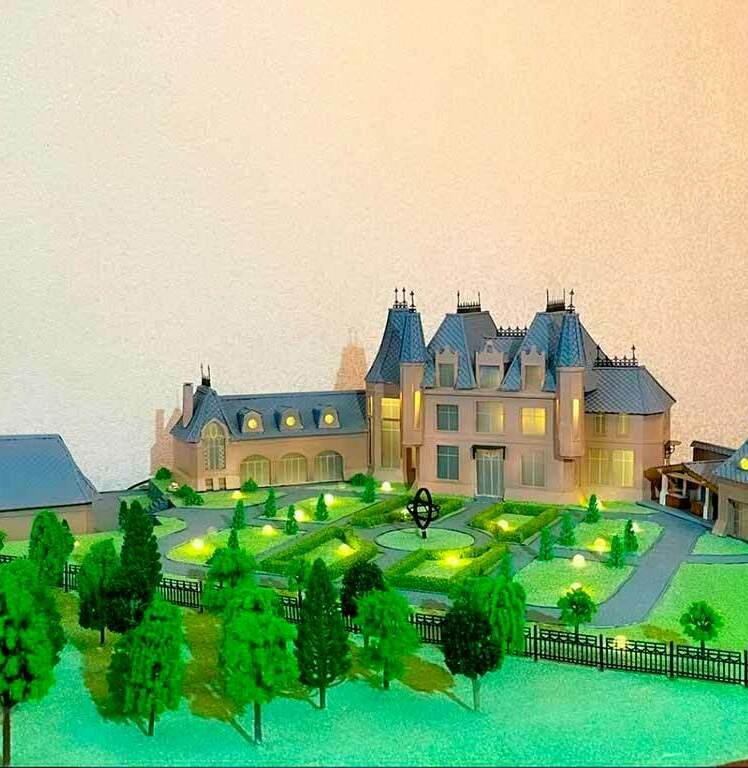 Масштабный макет роскошного особняка с французским ландшафтным дизайном — детализированная архитектурная модель для презентации элитных загородных резиденций Детализированная модель загородного особняка в стиле французского шато: трёхэтажное здание с башенками и мансардными окнами, окружённое ухоженным садом. На макете видны симметричные партерные клумбы, декоративные кустарники, стройные ряды деревьев, аккуратные дорожки и элементы освещения. Присутствуют хозяйственные постройки, гармонично вписанные в общий архитектурный ансамбль. Модель демонстрирует продуманную планировку территории, сочетание классической архитектуры и изысканного ландшафтного дизайна — идеальный пример для визуализации проектов элитных загородных домов и планирования садово-парковых зон.