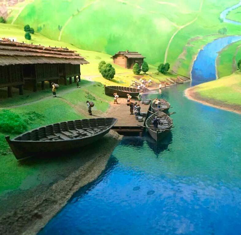 Макет изготовленный для музея на заказ тщательно выполненный макет древнего или старинного речного поселения. В центре композиции деревянный причал, к которому пришвартованы несколько лодок. На пристани и у берега стоят крошечные фигурки людей: кто‑то выгружает товар, кто‑то собирается в путь по реке. Слева вытянулось вдоль берега длинное дощатое строение на сваях — словно склад или амбар, под которым можно спрятаться от солнца и дождя. По тропинкам между травяными холмами движутся люди с ношей за спиной. Вдали, на пологих зелёных склонах, видны ещё постройки и стадо животных. Ярко‑синяя река, изгибаясь, уходит в глубь макета, растворяясь среди холмов и придавая всей сцене ощущение спокойной, размеренной жизни у водного пути.