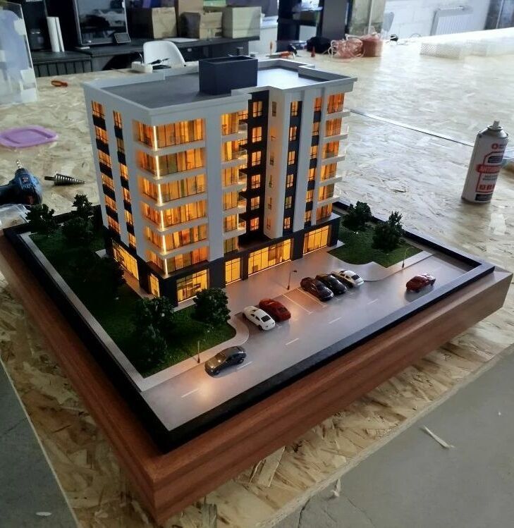3D-масштабный макет современного многоэтажного жилого здания с подсветкой фасадов. Модель демонстрирует планировку комплекса: главный корпус с балконами и большими окнами, входная группа, крытая парковка с несколькими автомобилями, благоустроенные зелёные зоны с деревьями и кустарниками по периметру. Видны элементы городской инфраструктуры — проезжая часть с разметкой, тротуар, уличные фонари. Макет выполнен с высокой детализацией, включая текстуры фасадов, ландшафт и миниатюрные автомобили, что позволяет наглядно представить будущий объект и его интеграцию в городскую среду. Идеален для презентации девелоперских проектов инвесторам и будущим жильцам.
