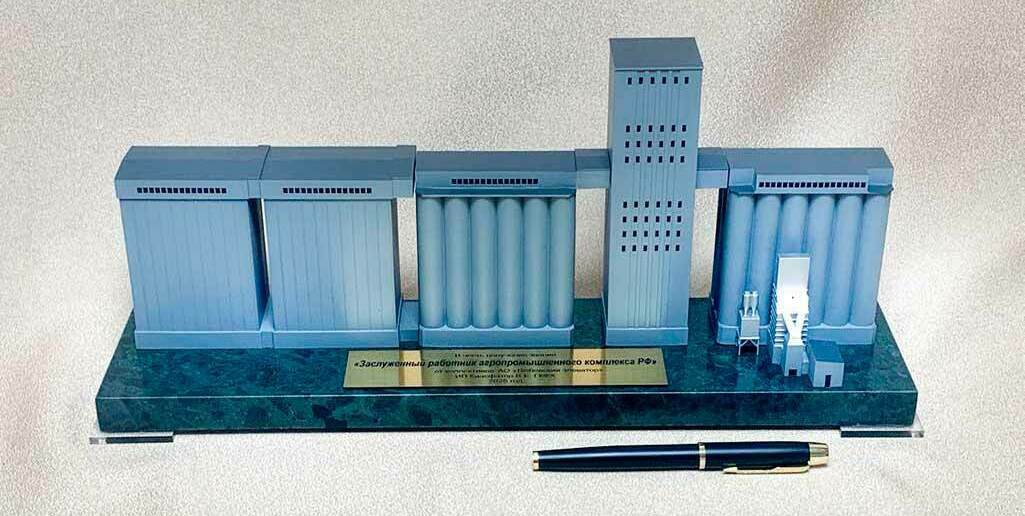 Несколько корпусов хранения: блоки цилиндрических силосов (силосные батареи) и прямоугольные секции складов. • Центральная рабочая (норийная) башня с окнами — узел вертикального транспортирования зерна (нории, распределение по силосам). • Надсилосные и межкорпусные галереи — закрытые конвейерные переходы между секциями. • Узлы приёма/подготовки зерна у правого блока (оборудование очистки/аспирации, загрузочные устройства). • Основание‑подставка с табличкой (памятный/наградной макет для работников АПК).  Назначение объекта: приём, транспортирование, очистка и распределение зерна по силосам для длительного хранения и последующей отгрузки.