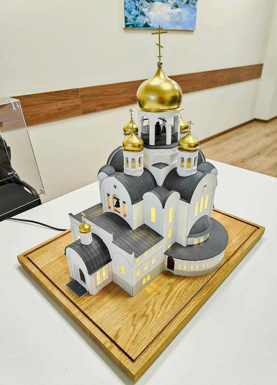 Макет православного храма с золотыми куполами: детализированная архитектурная модель для выставок и презентаций Детализированный масштабный макет православного храма: белокаменные стены, арочные окна и порталы, чёрные кровли, золотые купола с крестами; в проёмах — тёплая подсветка, у входа — тёмная площадка с крапчатой фактурой.