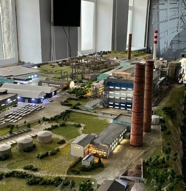 Детализированный 3D-масштабный макет промышленного предприятия / завода, расположенного на берегу водоёма. В композиции — крупный производственный корпус с конвейерными линиями и технологическим оборудованием, несколько вспомогательных цехов и складских помещений. Выделяются две высокие дымовые трубы, одна из которых окрашена в красно-белые полосы. Слева размещён грузовой порт с причалом, кранами для погрузки/разгрузки и пришвартованным судном. Видны транспортировочные пути, подъездные дороги, элементы ландшафтного дизайна (склоны, растительность). Макет демонстрирует полный цикл работы промышленного комплекса — от приёма сырья через производственные процессы до отгрузки готовой продукции. Реалистичная имитация инфраструктуры с высокой степенью детализации для использования в презентациях, планировании и визуализации проектов.