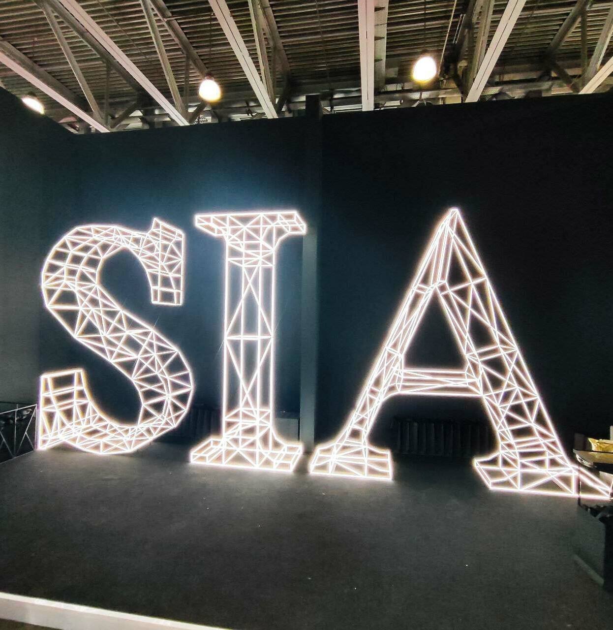 3‑D масштабный макет объёмных букв SIA: белая полигональная сетка, яркая светодиодная подсветка, чёткие грани и каркасная структура; индустриальный стиль, тёмный фон, зона отдыха с мебелью, акцент на современном оформлении выставочных и коммерческих площадок3‑D масштабный макет букв SIA: полигональная каркасная структура с равномерной светодиодной подсветкой, белый контур, чёткая геометрия граней; композиция размещена в тёмном пространстве с индустриальным потолком, справа — зона с креслами, слева — экран.