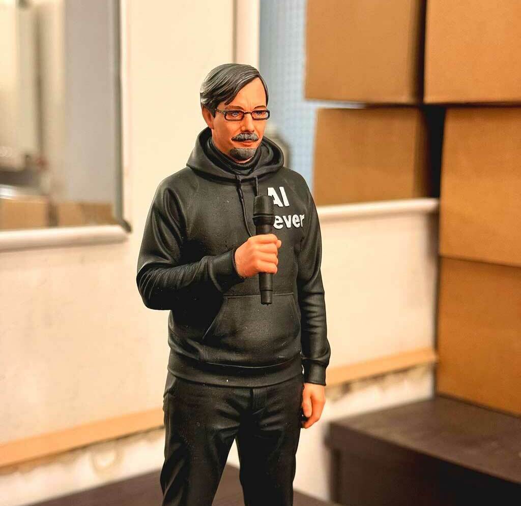 Реалистичная 3D-фигурка человека на заказ по фото: детализированная статуэтка с микрофоном, напечатанная на 3D-принтере. Кастомная модель в чёрной худи с надписью — идеальный сувенир для коллекционеров и поклонников публичных выступлений! 3D-фигурка мужчины, изготовленная по фотографии на 3D-принтере. Статуэтка изображает человека в расслабленной позе с микрофоном в руке. Одет в чёрную худи с надписью «AI ever», чёрные брюки, носит очки и аккуратную бороду. Высокая детализация: проработаны черты лица, текстура одежды, поза передаёт атмосферу публичного выступления. Фигурка отлично подойдёт для украшения офиса, в качестве оригинального подарка или элемента коллекции 3D-моделей. Пример персонализированной печати с сохранением индивидуальных особенностей.