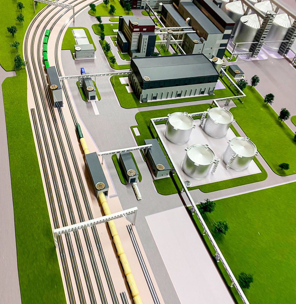 Детализированный макет промышленного предприятия: видны производственные корпуса, резервуары для хранения сырья, складские помещения, разветвлённая сеть железнодорожных путей с грузовыми составами, погрузочно-разгрузочные узлы. Модель демонстрирует взаимосвязь технологических зон, логистику перемещения грузов, инженерные коммуникации и планировку территории с озеленением. Реалистичная проработка элементов — от эстакад до малых архитектурных форм — делает макет идеальным для выставок, презентаций инвестиционных проектов и образовательных целей.