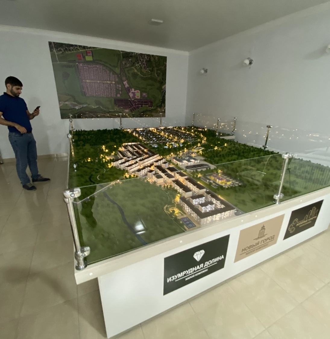 Масштабный архитектурный макет коттеджного посёлка «Изумрудная долина» — выставочная модель, изготовленная на заказ в Студии макетов: детальная визуализация жилого комплекса с инфраструктурой Детально проработанный архитектурный макет жилого комплекса «Изумрудная долина» (категория «Новый город») с имитацией вечерней подсветки. Включает макеты многоэтажных и малоэтажных зданий, благоустроенных зон отдыха, парковых территорий, подъездных путей и элементов инфраструктуры. Установлен на выставочном стенде с защитным стеклом, дополнен крупномасштабной схемой застройки на стене. Модель демонстрирует планировку посёлка, распределение жилых и общественных зон, концепцию озеленения и освещения. Создана на заказ для презентации проекта на выставках и встречах с инвесторами.