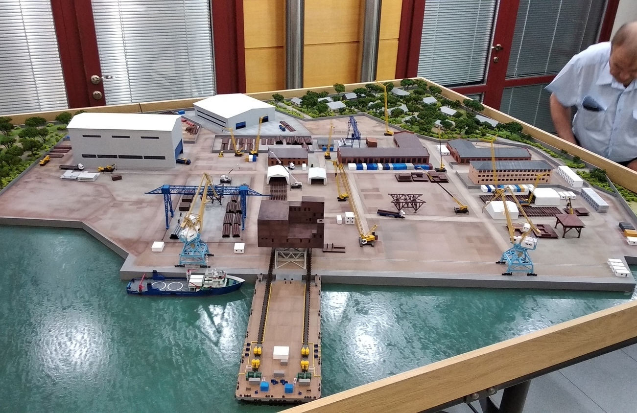 3D масштабный макет морского порта с инфраструктурой: детализированная модель производственной площадки, причального фронта, грузовых кранов и судна для презентации логистических проектов инвесторам и заказчикам Масштабный 3D макет морского порта, демонстрирующий комплексную промышленную площадку. В композиции — причальный фронт с пришвартованным грузовым судном, сеть подъездных путей, складские и производственные здания. Реализована развитая портовая инфраструктура: несколько портальных и башенных кранов синего цвета, погрузочная техника, контейнеры, зоны складирования. Водная поверхность имитирует акваторию с реалистичной текстурой. На территории макета проработаны элементы благоустройства — зеленые насаждения по периметру. Макет выполнен с высокой детализацией для наглядной демонстрации структуры и логистики портового комплекса на выставках и презентациях.