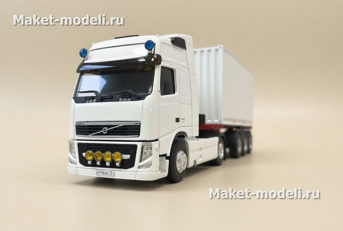 Масштабная модель магистрального седельного тягача Volvo FH 480, колесная формула 4×2, с трехосным полуприцепом-фургоном (контейнеровозом) Тягач компоновки с высокой спальной кабиной (high-roof sleeper cab). • Передняя светотехника: основные фары, блок дополнительных противотуманных/дальнобойных фар на переднем бампере. • Солнечный козырёк над ветровым стеклом, сигнальные огни на крыше кабины. • Фирменная решётка радиатора Volvo с логотипом и обозначением модели FH и мощности «480». • Смоделированные элементы ходовой части: односкатные шины на управляемой передней оси и ведущей задней. • Сцепка с трёхосным полуприцепом прямоугольного сечения (контейнер/фургон). Модель установлена на ровной поверхности, на фоне стеллажей с ячейками для хранения и коробок с другими моделями.