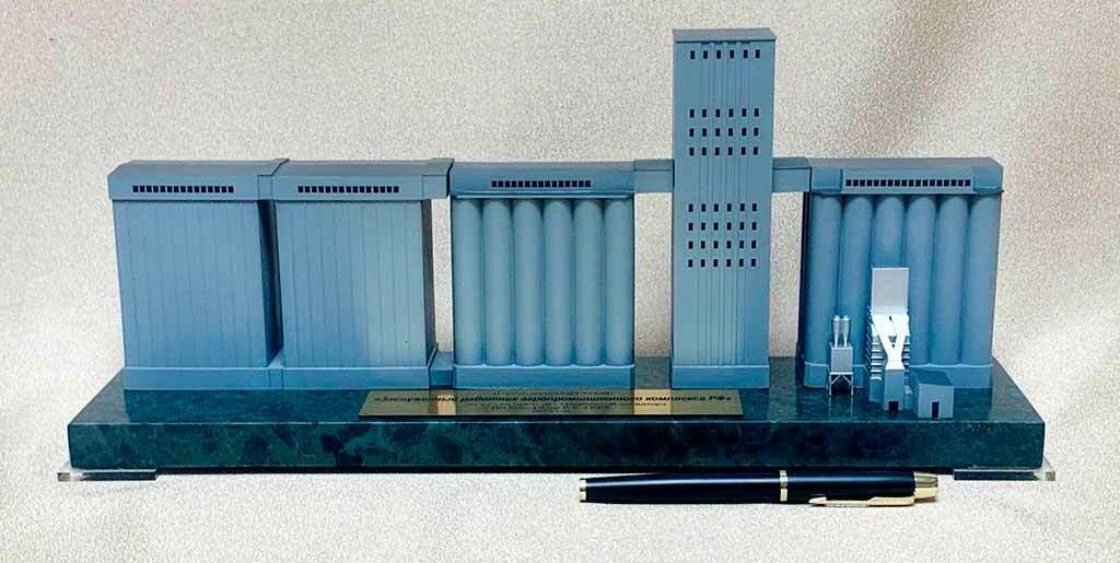 На каменной подставке установлен однотонный серо‑голубой макет промышленного зернохранилища. • Слева и справа — корпуса и батареи цилиндрических силосов, соединённые надсилосными галереями. • В центре — высокая норийная (элеваторная) башня с рядами технологических проёмов. • Справа у силосов показаны мелкие узлы: оборудование приёмки/очистки и загрузки. • Спереди лежит ручка, подчёркивая настольный формат и масштаб модели. Смысл: демонстрационный или подарочный макет типового зернохранилищного комплекса (элеватора).