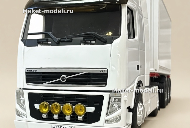 Масштабная модель магистрального седельного тягача Volvo FH 480, колесная формула 4×2, с трехосным полуприцепом-фургоном (контейнеровозом) Тягач компоновки с высокой спальной кабиной (high-roof sleeper cab). • Передняя светотехника: основные фары, блок дополнительных противотуманных/дальнобойных фар на переднем бампере. • Солнечный козырёк над ветровым стеклом, сигнальные огни на крыше кабины. • Фирменная решётка радиатора Volvo с логотипом и обозначением модели FH и мощности «480». • Смоделированные элементы ходовой части: односкатные шины на управляемой передней оси и ведущей задней. • Сцепка с трёхосным полуприцепом прямоугольного сечения (контейнер/фургон). Модель установлена на ровной поверхности, на фоне стеллажей с ячейками для хранения и коробок с другими моделями.