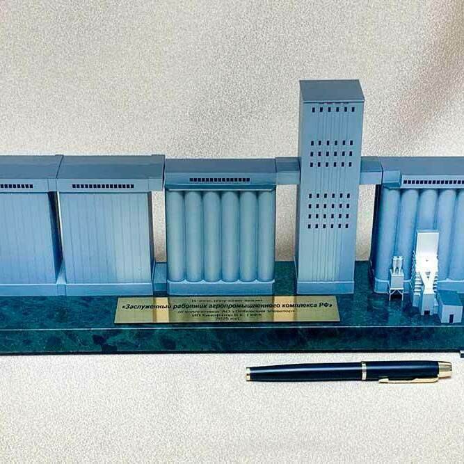 3D-масштабный макет зернового элеватора, демонстрирующий ключевые элементы технологического комплекса: три прямоугольных блока модульных силосов для хранения зерна, центральная группа вертикальных цилиндров (силосные батареи) под общей крышей-галереей, высокая прямоугольная норийно-распределительная башня с технологическими площадками. Справа расположен миниатюрный приёмно-очистительный узел с загрузочной эстакадой и аспирационными фильтрами. Макет установлен на презентационном постаменте с табличкой, рядом лежит ручка для демонстрации масштаба. Детализированная модель наглядно иллюстрирует процессы приёма, очистки, хранения и отгрузки зерна, подходит для использования в выставочных и образовательных целях.