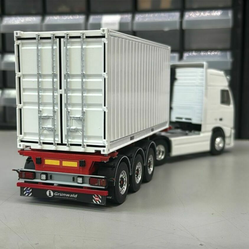 Масштабная модель магистрального тягача-автопоезда: Что видно на фото (сзади): • Задняя часть трёхосного полуприцепа с коробчатым кузовом. • Двустворчатые задние двери с вертикальными запорными штангами и петлями. • Задний защитный брус (RUPD) с фонарями и светоотражателями. • Трёхосная тележка полуприцепа с односкатной ошиновкой. • Грязезащитные щитки с логотипом Grünwald. • В глубине кадра — седельный тягач Volvo белого цвета с высокой кабинной надстройкой, состыкованный с полуприцепом через седельно-сцепное устройство.