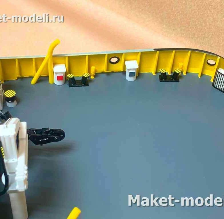  Серый настил рабочей палубы; по периметру — бульварк (внутренний борт) жёлтого цвета с продольными и поперечными ребрами жёсткости. • Швартовая арматура: одиночные тумбы/кнехты и парные битенги, отмеченные чёрно‑жёлтой сигнальной разметкой. • Роликовый клюз/направляющий ролик для швартовного либо буксирного троса. • Шкафы и боксы для противопожарного и аварийно‑спасательного имущества (ящик с рукавом/огнетушителем, кнопка/пост сигнализации). • Корзина/контейнер для хранения тросов у борта. • Круглые отверстия в бульварке (шпигаты/дренаж) и леерное ограждение с трапом у надстройки слева.  Назначение макета: демонстрация планировки и компоновки швартовно‑буксирного и противопожарного оснащения на рабочей палубе технического судна.