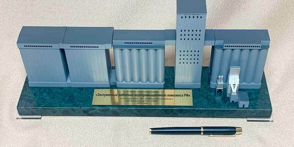 На каменном основании стоят серые модули: слева и справа — блоки вертикальных цилиндрических силосов, в центре — высокая прямоугольная башня-элеватор (головная), между секциями — галереи-конвейеры. Справа видны небольшие узлы перегрузки/очистки. На табличке — надпись «Заслуженный работник агропромышленного комплекса РФ», что указывает на памятно-наградное назначение макета. Для масштаба спереди лежит ручка.