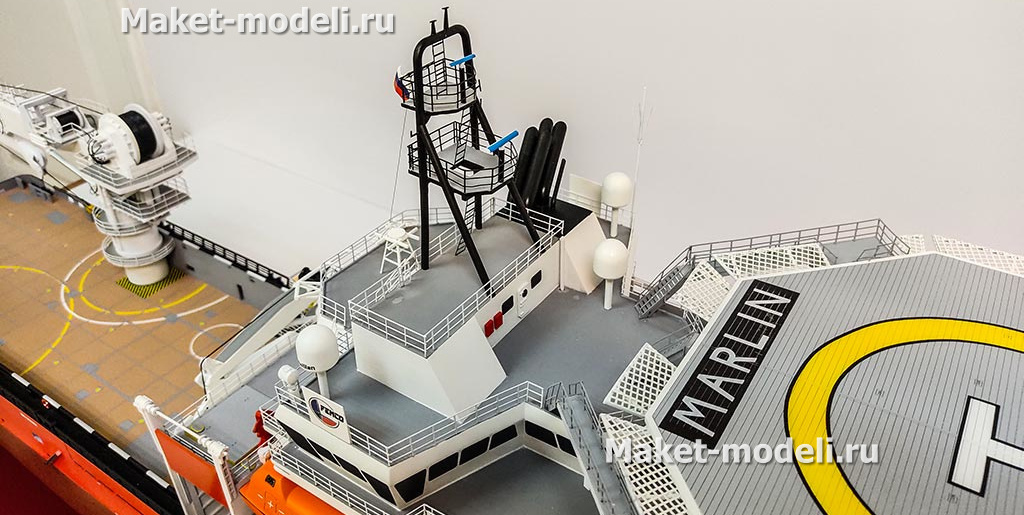 3‑D масштабная модель судна обеспечения: красный корпус, белая надстройка, детализированные винторулевые колонки и палубное оборудование»Перед нами 3‑D масштабная модель судна обеспечения — высокодетализированная копия с красным корпусом, белой надстройкой, палубными кранами, спасательными шлюпками и винтами; экспонат размещён на опорах в выставочном пространстве.