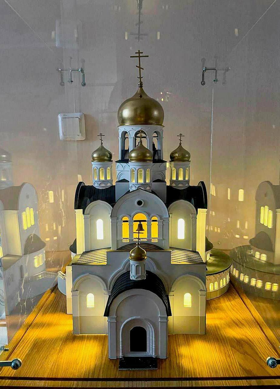 Макет православного храма с золотыми куполами: детализированная архитектурная модель для выставок и презентаций Детализированный масштабный макет православного храма: белокаменные стены, арочные окна и порталы, чёрные кровли, золотые купола с крестами; в проёмах — тёплая подсветка, у входа — тёмная площадка с крапчатой фактурой.