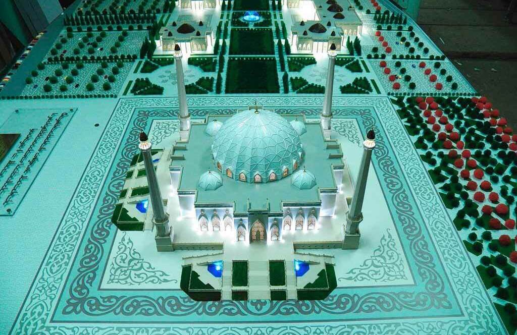 Детальный масштабный макет архитектурного комплекса мечети: в центре — здание с крупным геодезическим куполом, окружённое четырьмя высокими минаретами. Фасад мечети украшен ажурные арочными входами с внутренней подсветкой, декоративными решётками и орнаментальной плиткой. Перед мечетью — широкие ступени, ведущие к главному входу. Территория вокруг здания тщательно проработана: симметричные аллеи с миниатюрными деревьями, зелёные газоны, синие декоративные бассейны с подсветкой. На заднем плане — дополнительные павильоны и архитектурные элементы комплекса, выполненные с высокой детализацией. Модель демонстрирует гармонию исламской архитектуры и ландшафтного дизайна, идеально подходит для презентации градостроительных проектов и выставочных экспозиций.