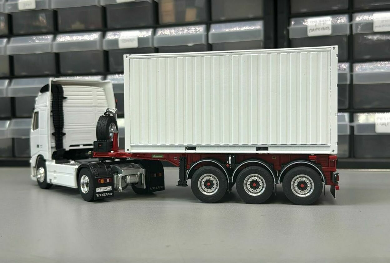  белый седельный тягач Volvo с высокими кабиной и задней стенкой,   • седельно-сцепное устройство (седло) на раме тягача,   • смонтированное на полуприцепе запасное колесо,   • трёхосный полуприцеп красного цвета,   • установленный на полуприцепе белый прямоугольный контейнер (имитация стандартного морского/интермодального контейнера).   Фон — стеллаж с пластиковыми ячейками, что подчёркивает, что это именно коллекционная/масштабная модель, а не реальный автопоезд.