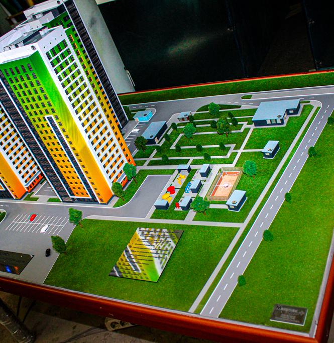 Масштабный архитектурный макет современного жилого комплекса с высотными башнями и развитой инфраструктурой — детализированная выставочная модель, изготовленная на заказ в Студии макетов. Визуализация жилых зданий, парковых зон, детских площадок и дорожной сети в едином ансамбле Масштабная модель жилого комплекса: две высотные башни с яркой цветовой отделкой фасадов (зелёные и оранжевые акценты), окружённые благоустроенной территорией. В композиции представлены элементы городской инфраструктуры — асфальтированные дороги с разметкой, парковочные зоны, пешеходные дорожки. Проработаны зоны отдыха: детская игровая площадка с безопасным покрытием, спортивная зона, зелёные насаждения с миниатюрными деревьями и кустарниками. Модель демонстрирует концепцию комфортной городской среды, сочетающей современную архитектуру с продуманным ландшафтным дизайном. Идеальна для презентации девелоперских проектов на выставках, архитектурных конкурсах и встречах с инвесторами. Выполнена на заказ в Студии макетов с соблюдением точных пропорций и масштаба.