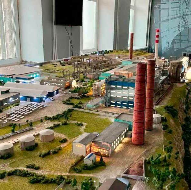 Масштабный макет промышленного комплекса: центральное место занимают высокие дымовые трубы красно-белой окраски, символизирующие энергетическое производство. Вокруг расположены производственные здания с синими и серыми фасадами, складские помещения и административные корпуса. Видны резервуары для хранения сырья или готовой продукции, аккуратно размещённые на зелёной территории. Проработаны подъездные пути, железнодорожные линии и зоны погрузки/разгрузки. На макете также изображены элементы ландшафтного дизайна — миниатюрные деревья, кустарники, газоны. Справа заметен участок с горнодобывающим оборудованием и откосами, имитирующими карьер. Модель демонстрирует планировку комплекса, взаимосвязь зданий и инфраструктурных объектов, позволяет оценить масштаб и логистику предприятия. Подходит для визуализации проекта, презентации инвесторам и использования в архитектурных выставках.