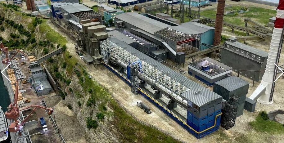 Детализированный 3D-масштабный макет промышленного предприятия / завода, расположенного на берегу водоёма. В композиции — крупный производственный корпус с конвейерными линиями и технологическим оборудованием, несколько вспомогательных цехов и складских помещений. Выделяются две высокие дымовые трубы, одна из которых окрашена в красно-белые полосы. Слева размещён грузовой порт с причалом, кранами для погрузки/разгрузки и пришвартованным судном. Видны транспортировочные пути, подъездные дороги, элементы ландшафтного дизайна (склоны, растительность). Макет демонстрирует полный цикл работы промышленного комплекса — от приёма сырья через производственные процессы до отгрузки готовой продукции. Реалистичная имитация инфраструктуры с высокой степенью детализации для использования в презентациях, планировании и визуализации проектов.