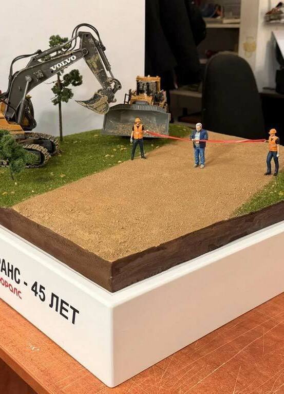 3D-фигурка человека на заказ по фото: реалистичная миниатюра с церемонией перерезания ленты. Кастомная модель в джинсах и стёганой куртке с детализацией одежды и мимики, напечатанная на 3D-принтере для диорамы стройплощадки. Уникальный сувенир для коллекционеров! Реалистичная 3D-фигурка человека, изготовленная по фотографии на 3D-принтере, изображает мужчину в момент перерезания красной ленты — символического акта открытия. Фигурка выполнена с высокой детализацией: мужчина в синей стёганой куртке, светло-зелёной футболке и синих джинсах, с аккуратной сединой и выразительным лицом. В руках держит ножницы, готовясь перерезать ленту. Рядом стоит фигурка строителя в оранжевом жилете и каске, наблюдающего за церемонией. На заднем плане — детализированная модель строительной техники (бульдозер). Композиция идеально подходит для диорамы стройплощадки, коллекционных экспозиций или в качестве оригинального подарка. Пример персонализированной 3D-печати с передачей индивидуальных особенностей и атмосферы события.