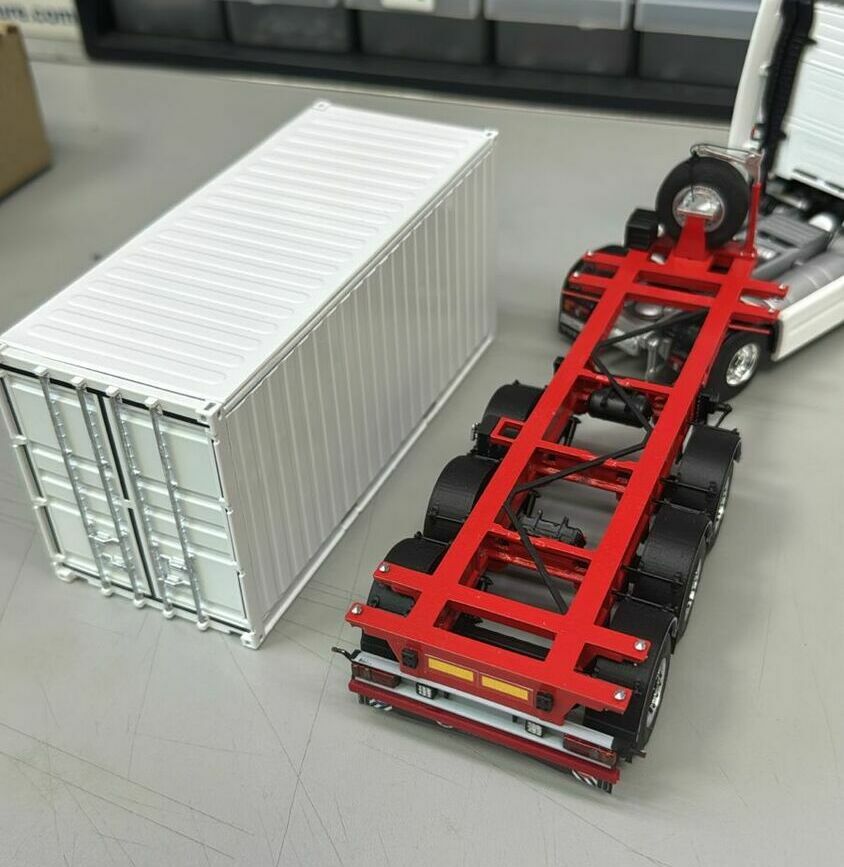  Седельный тягач (частично виден справа) с пятой точкой сцепления.   • Трёхосный контейнерный полуприцеп *скелетного типа* (frame / skeletal semi‑trailer) с красной рамой, предназначенный для перевозки морских контейнеров.   • Отдельно стоящий универсальный сухогрузный ISO‑контейнер (вероятнее всего 20‑футовый), с каркасом, продольной гофрой стенок и двустворчатыми запорными дверями.