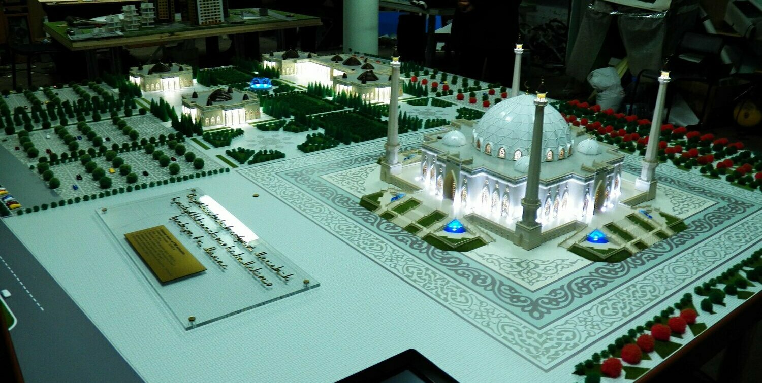 Масштабная модель белой мечети на выставочном подиуме: крупный геодезический купол, четыре высоких минарета со светодиодной подсветкой, ажурные арочные входы, ступенчатые террасы с синими декоративными фонтанами, орнаментальная площадь вокруг, симметричные зелёные насаждения и миниатюрные деревья, информационная табличка и сенсорный экран, высокий уровень проработки деталей