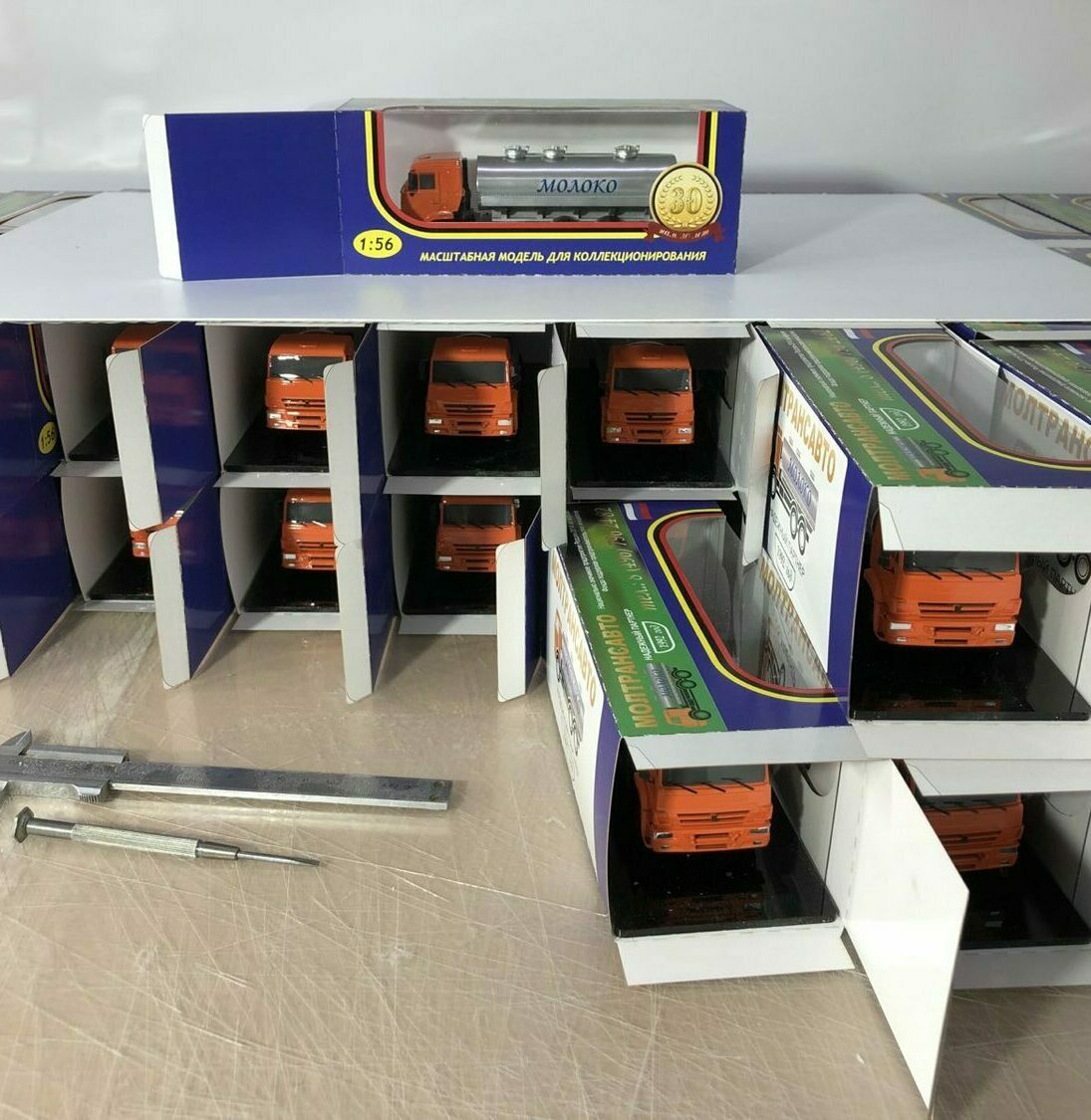 3D-масштабная модель грузовика-молоковоза с цистерной, подписанной «Молоко», в масштабе 1:56. Яркая оранжевая кабина и серебристая цистерна выполнены с высокой детализацией — видны решётка радиатора, зеркала заднего вида, колёса с проработанным протектором. Модель упакована в презентабельную коробку с прозрачным окном, предназначена для коллекционирования. Рядом расположены другие экземпляры в аналогичных боксах, создающие эффект выставочной экспозиции. В нижней части кадра — штангенциркуль и измерительный инструмент, подчёркивающие точность исполнения масштабной модели.