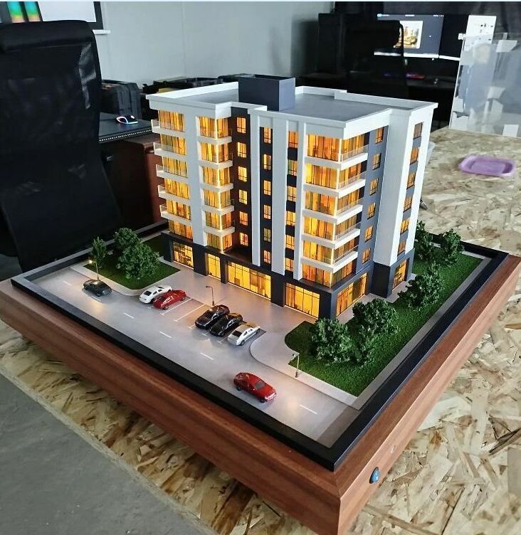 3D-масштабный макет современного многоэтажного жилого здания с подсветкой фасадов. Модель демонстрирует планировку комплекса: главный корпус с балконами и большими окнами, входная группа, крытая парковка с несколькими автомобилями, благоустроенные зелёные зоны с деревьями и кустарниками по периметру. Видны элементы городской инфраструктуры — проезжая часть с разметкой, тротуар, уличные фонари. Макет выполнен с высокой детализацией, включая текстуры фасадов, ландшафт и миниатюрные автомобили, что позволяет наглядно представить будущий объект и его интеграцию в городскую среду. Идеален для презентации девелоперских проектов инвесторам и будущим жильцам.