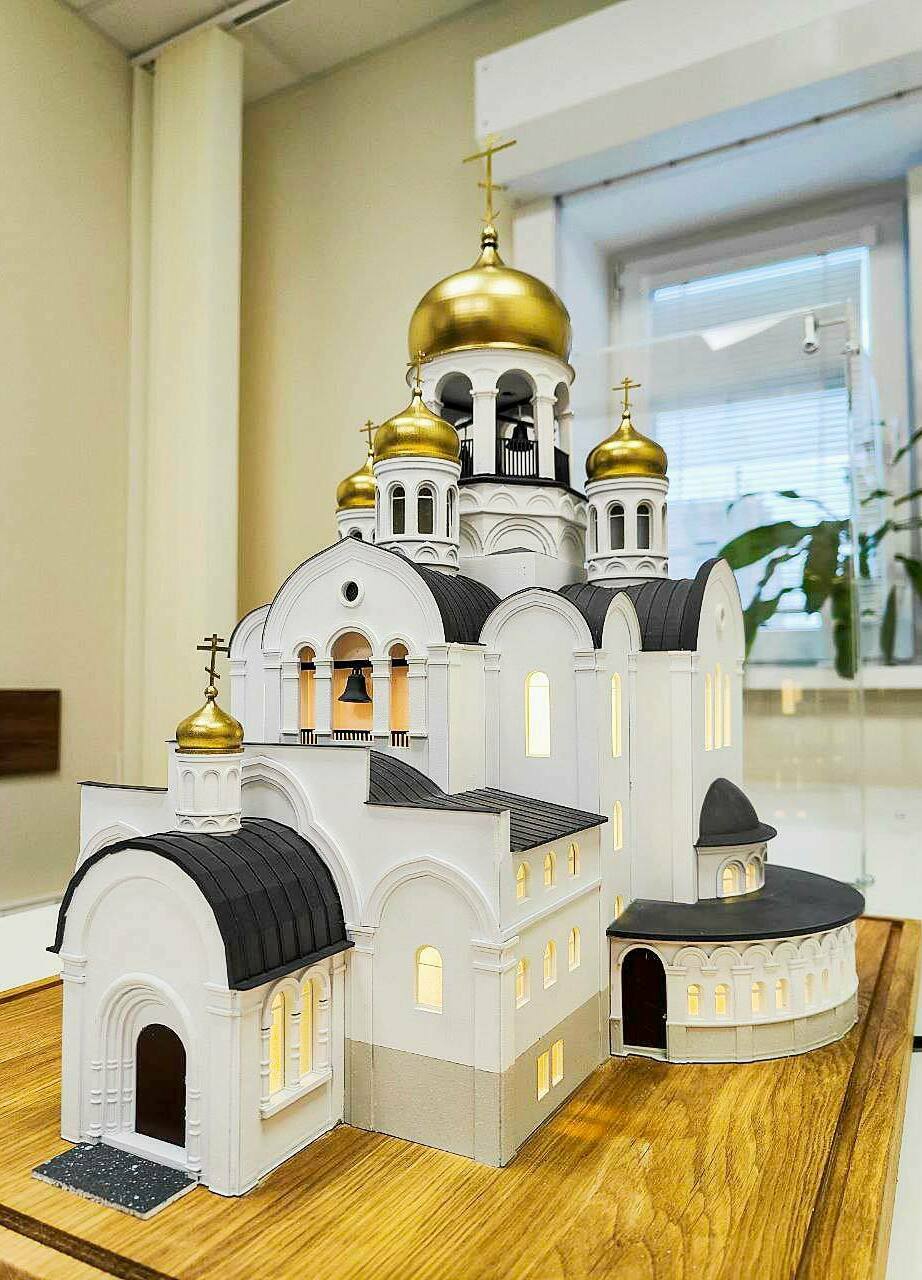 Макет православного храма с золотыми куполами: детализированная архитектурная модель для выставок и презентаций Детализированный масштабный макет православного храма: белокаменные стены, арочные окна и порталы, чёрные кровли, золотые купола с крестами; в проёмах — тёплая подсветка, у входа — тёмная площадка с крапчатой фактурой.
