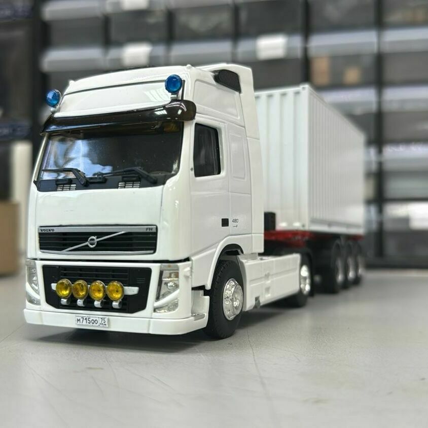 масштабная модель магистрального седельного тягача Volvo FH с полуприцепом.  Техническое описание кадра:   • Тягач Volvo FH, исполнение с высокой спальной кабиной (high roof sleeper cab), колёсная формула 4×2.   • На крыше кабины установлены сигнальные проблесковые маяки синего цвета; над ветровым стеклом — аэродинамический солнцезащитный козырёк.   • В переднем бампере смонтирован блок дополнительных противотуманных/дальнобойных фар (четыре круглых прожектора).   • Сцепное устройство типоразмера «седло» для работы с полуприцепом.   • Полуприцеп — трёхосный контейнерный полуприцеп (контейнеровоз) с установкой универсального крупнотоннажного контейнера.  