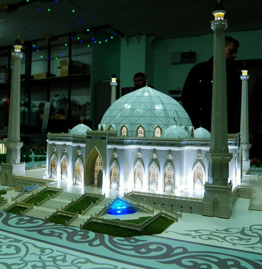Масштабный архитектурный макет белой мечети на подиуме: большой геодезический купол, четыре высоких минарета, светящиеся ажурные арочные ворота и окна, ступени, зелёные газоны и синие декоративные бассейны; интерьер мастерской на заднем плане изготовленный на заказ