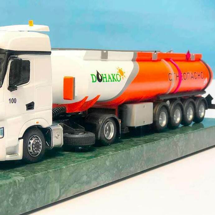 Масштабная модель бензовоза КАМАЗ с прицепом-цистерной DOHAKO в масштабе 1:43. Белая кабина с чёрными элементами и чётко видимым логотипом «KAMAZ», номер «100» на дверях. Оранжево-белая автоцистерна с логотипом DOHAKO и предупреждающей надписью «ОГНЕОПАСНО». Модель установлена на зелёной подставке, видны детали: фары, зеркала, колёса, подвеска, светоотражатели. Идеальна для выставок, посвящённых транспортной технике, и для коллекционеров масштабных моделей. Высокая детализация позволяет рассмотреть конструктивные особенности топливозаправщика.