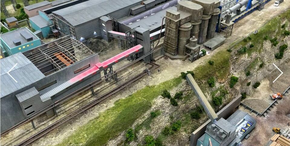 Детализированный 3D-масштабный макет промышленного предприятия / завода, расположенного на берегу водоёма. В композиции — крупный производственный корпус с конвейерными линиями и технологическим оборудованием, несколько вспомогательных цехов и складских помещений. Выделяются две высокие дымовые трубы, одна из которых окрашена в красно-белые полосы. Слева размещён грузовой порт с причалом, кранами для погрузки/разгрузки и пришвартованным судном. Видны транспортировочные пути, подъездные дороги, элементы ландшафтного дизайна (склоны, растительность). Макет демонстрирует полный цикл работы промышленного комплекса — от приёма сырья через производственные процессы до отгрузки готовой продукции. Реалистичная имитация инфраструктуры с высокой степенью детализации для использования в презентациях, планировании и визуализации проектов.